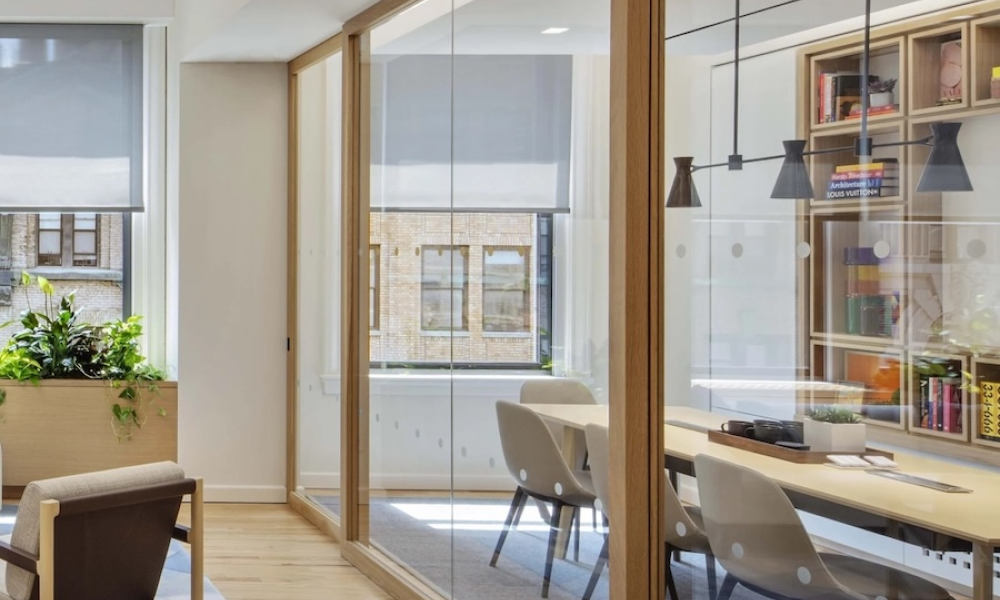Meeting room shown through floor-to-ceiling glass sliding doors. Automated shades are partially lowered in the background.